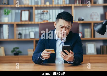 Verärgert Mann versucht, einen Kauf in einem Online-Shop zu machen, asiatische Geschäftsmann arbeitet in einem klassischen Büro, hält ein Telefon und eine Bankkreditkarte Stockfoto