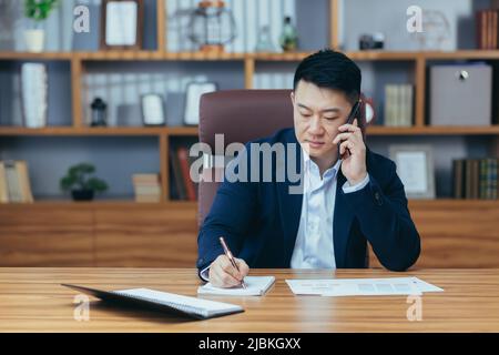 Workflow. Junger, gutaussehender, erfolgreicher asiatischer Mann, der im Büro arbeitet, das Telefon benutzt und am Telefon telefoniert. Direktor, Bankier, Anwalt, der an der TA sitzt Stockfoto