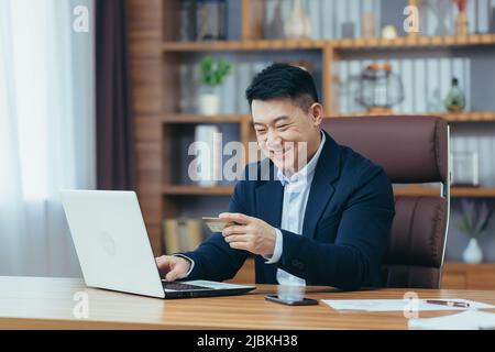 Der erfolgreiche asiatische Geschäftsmann führt Bankgeschäfte durch und überweist Geld von einem elektronischen Konto, verwendet einen Laptop und eine Bankkreditkarte Stockfoto