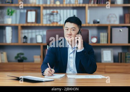 Workflow. Junger, gutaussehender, erfolgreicher asiatischer Mann, der im Büro arbeitet, das Telefon benutzt und am Telefon telefoniert. Direktor, Bankier, Anwalt, der an der TA sitzt Stockfoto