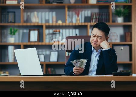 Unzufriedener asiatischer Chefunternehmer, der im Büro mit Bargeld in US-Dollar arbeitet, frustriert durch niedriges Einkommen Stockfoto