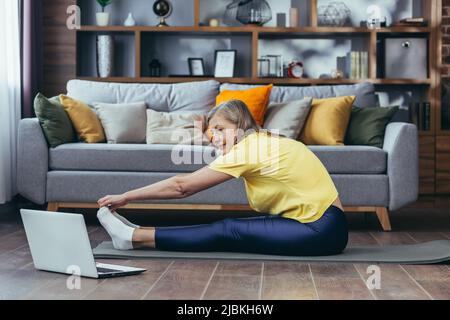 Ältere grauhaarige Frau, die zu Hause Fitness macht, auf einer Matte sitzt, Online-Aerobic-Kurse macht, Stretching macht, auf den Laptop-Bildschirm schaut Stockfoto