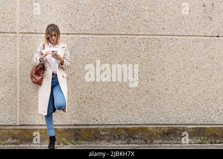 Frau in beigem Mantel, weißem Hemd und Jeans, die an der Wand gelehnt sind und das Handy betrachten Stockfoto