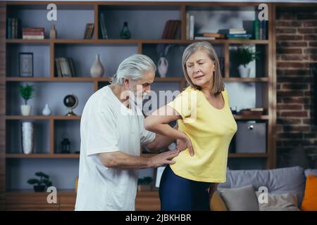 Der ältere Ehemann massierte während des Fitness-Kurses zu Hause den Rücken seiner Frau und das grauhaarige Rentnerpaar Stockfoto