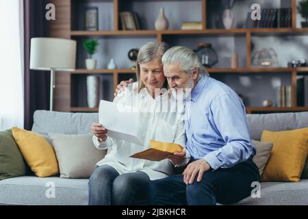 Glückliches Rentnerpaar, das zu Hause auf dem Sofa sitzt und den Brief liest, glücklich und lächelnd Stockfoto