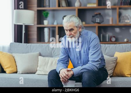 Der ältere grauhaarige Mann sitzt zu Hause auf dem Sofa, nachdem er ein stumpfes Blatt gewonnen hat, in Depressionen Stockfoto