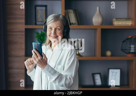 Eine ältere moderne Frau zu Hause am Fenster hört Musik am Telefon, nutzt eine Musikanwendung für das Online-Hören und Kopfhörer Stockfoto