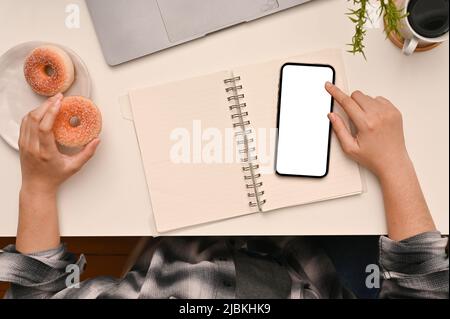 Die Topaufnahme einer College-Studentin im Scott-Hemd sitzt in ihrem modernen Arbeitsplatz, indem sie Smartphone und Donut isst. Stockfoto