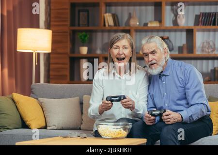 Ältere Paare spielen Videospielkonsole mit Joysticks sitzen auf der Couch im Wohnzimmer. Lustige ältere Menschen Familie mit Spaß verbringen Freizeit toge Stockfoto