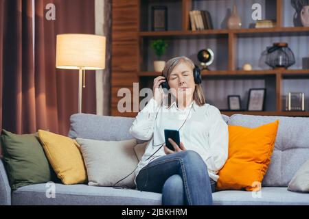 Ältere Frau, die sich abends zu Hause entspannt, ein Hörbuch und Podcast hört, die App auf ihrem Telefon und Kopfhörer verwendet Stockfoto