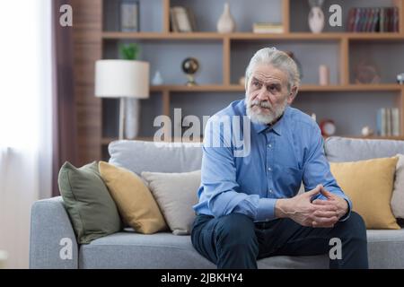 Älterer nachdenklicher Mann, der auf dem Sofa sitzt, einsamer grauhaariger Mann im Wohnzimmer, deprimiert Stockfoto