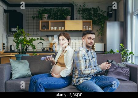 Ignorieren. Junges Paar, Schwangere und Mann sitzen zu Hause mit dem Rücken auf dem Sofa und halten Mobiltelefone in den Händen. Reden Sie nicht Stockfoto