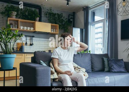 Eine junge Schwangere sitzt zu Hause auf der Couch, hat eine laufende Nase und Allergien, niest mit einer Serviette Stockfoto