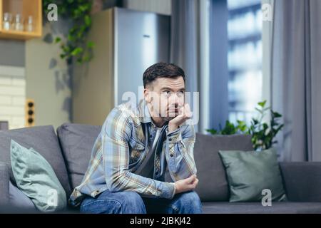 Porträt eines hübschen jungen Mannes. Warten, gelangweilt, gedacht. Sitzt zu Hause auf der Couch, hält seinen Kopf in der Hand, denkt an Depression, Quarantäne Stockfoto
