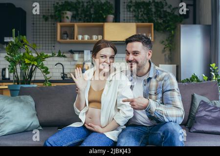 Junge Familie sitzt zu Hause auf dem Sofa. Sie sprechen bei einem Videoanruf. Schwanger Frau und Mann. Frau winkte mit der Hand zur Kamera und lächelte. Der Mann zeigt Stockfoto