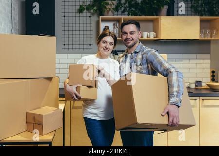 Porträt eines jungen Familienmannes und einer Schwangeren Frau in einer neuen Wohnung, die Kartons in der Hand hält und die Kamera anschaut und lächelt Stockfoto