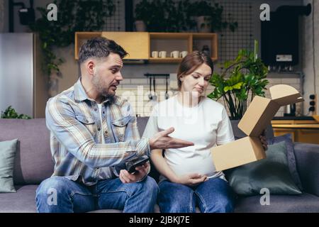 Eine junge Familie, eine Schwangere und ein Mann haben versehentlich das Paket einer anderen Person erhalten. Sie öffneten die Kiste, enttäuscht. Zu Hause auf dem Sofa sitzen Stockfoto