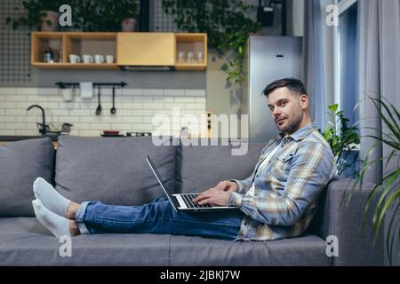 Freiberufler. Hübscher junger Mann, der zu Hause am Laptop arbeitet. Auf der Couch in Hauskleidung liegen. Stockfoto