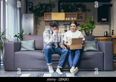 Eine junge Familie, eine Schwangere und ein Mann haben versehentlich das Paket einer anderen Person erhalten. Sie öffneten die Kiste, enttäuscht. Zu Hause auf dem Sofa sitzen Stockfoto