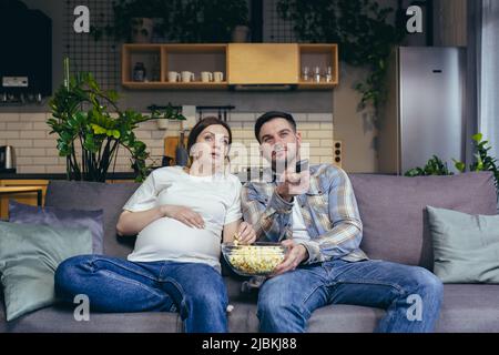 Heimkino. Junge Familie. Schwanger Frau und Mann sitzen zu Hause auf dem Sofa, schauen Fernsehen, Film, essen Popcorn. Viel Vergnügen miteinander Stockfoto