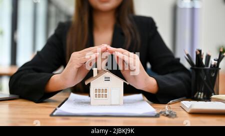 Beschnittenes Bild, weibliche Hausversichererin sitzt am Schreibtisch und macht eine Geste mit schützenden Händen über dem Haus. Sachversicherung und Sicherheit Stockfoto