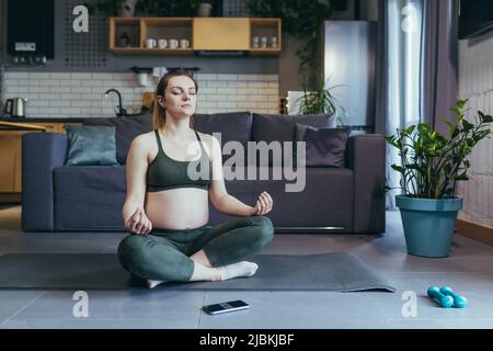 Eine Schwangerin meditiert und praktiziert Yoga in einer Lotusposition zu Hause Stockfoto