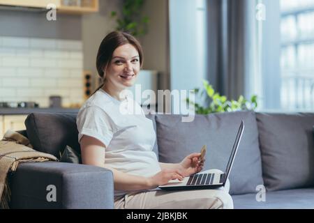 Eine Schwangerin sitzt zu Hause auf dem Sofa, kauft mit einem Laptop im Online-Shop ein, blickt auf die Kamera und hält lächelnd die Kreditkarte Stockfoto