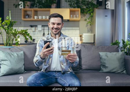 Porträt eines jungen, gutaussehenden Mannes. Verfügt über ein Telefon und eine Kreditkarte. Sitzt zu Hause auf der Couch, schaut auf das Zimmer, lächelt Stockfoto