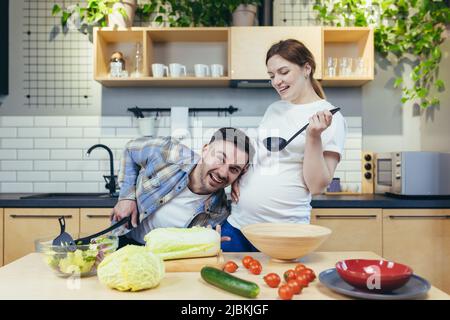 Eine junge Familie, eine Schwangere und ein Mann bereiten gemeinsam in der Küche gesundes und leckeres Essen zu. Viel Spaß zusammen. Der Mann wird seinem wi helfen Stockfoto