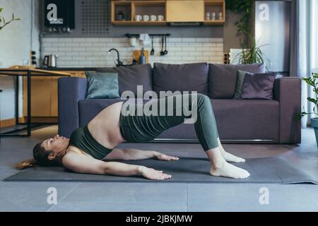 Eine Schwangere schöne Frau geht zu Hause zum Sport, sitzt und liegt in einer Sportuniform auf einer Sportmatte, macht Übungen, ist zufrieden Stockfoto