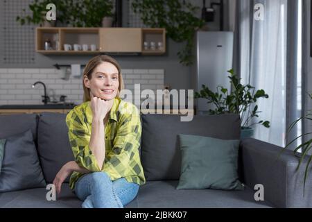 Porträt einer erfolgreichen Frau mittleren Alters, einer Hausfrau, die in der Küche auf dem Sofa sitzt und lächelt und die Kamera anschaut Stockfoto