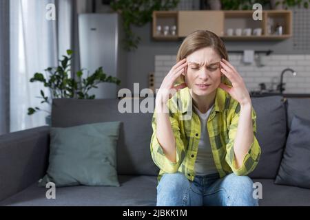Die Frau ist allein zu Hause, sitzt auf der Couch in der Küche, hat starke Kopfschmerzen Stockfoto