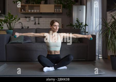 Eine Frau mittleren Alters sitzt auf dem Boden und führt Übungen mit Hanteln durch Stockfoto