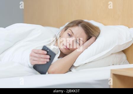 Eine Frau liegt morgens zu Hause unter einer Decke, schaut auf den Telefonbildschirm, liest die Morgennachrichten Stockfoto