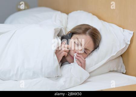 Nahaufnahme einer kranken Frau zu Hause, hat eine schlechte laufende Nase und Kälte, liegt mit einer Decke bedeckt im Bett, ruft einen Arzt um Rat Stockfoto