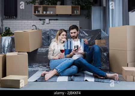 Ein verliebtes Ehepaar, ein Mann und eine Frau feiern eine Hauswarmfeier, trinken Wein aus Kartons und wählen neue Möbel und eine Haushalts-App Stockfoto