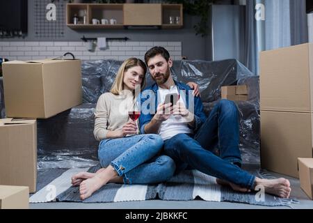 Ein verliebtes Ehepaar, ein Mann und eine Frau feiern eine Hauswarmfeier, trinken Wein aus Kartons und wählen neue Möbel und eine Haushalts-App Stockfoto