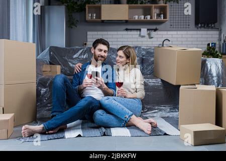 Ein verliebtes Ehepaar, ein Mann und eine Frau feiern eine Hauswarmfeier, trinken Wein aus Kartons und wählen neue Möbel und eine Haushalts-App Stockfoto