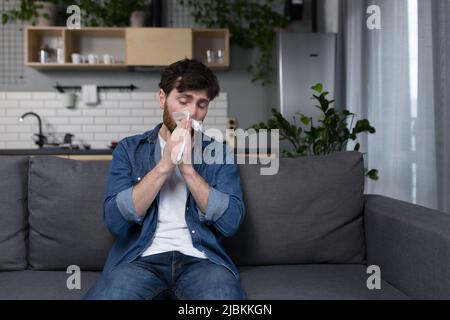 Der Mann ist zu Hause krank, hat Grippe und laufende Nase, niest, sitzt allein in der Küche auf der Couch Stockfoto