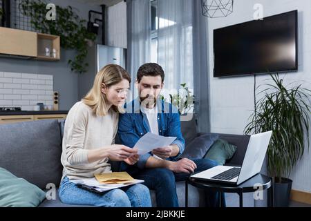 Ehemann und Ehefrau, junge Familie, die gemeinsam an den Haushaltsfinanzen arbeiten, über Rechnungen streiten, Laptop benutzen und zu Hause auf dem Sofa sitzen Stockfoto