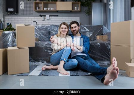 Glückliches junges Paar, Mann und Frau zu Hause, sitzen auf dem Sofa in der neu gemieteten Wohnung, in der Nähe von Kartons Stockfoto