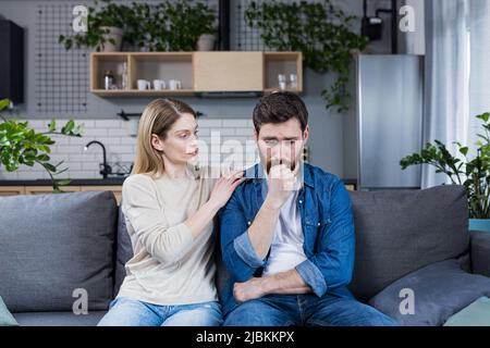 Eine Frau unterstützt einen verärgerten Ehemann, der versagt hat, ein Ehepaar sitzt auf der Couch Stockfoto