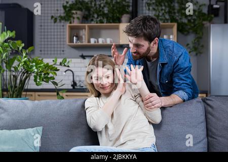 Häusliche Gewalt. Familienkonflikt. Ein Mann schreit und schlägt eine Frau zu Hause. Verängstigte Frau schließt ihre Arme und sitzt auf der Couch. Sie hat Angst Stockfoto