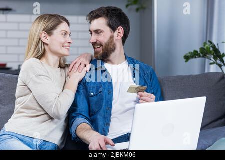 Online-Shopping. Familie, Mann und Frau zu Hause, sitzen auf der Couch, kaufen Waren online von einem Laptop, halten eine Kreditkarte in den Händen. Glücklich, Stockfoto