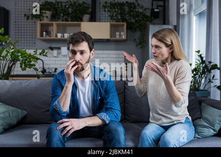 Familie streitet, Mann und Frau sitzen zu Hause auf dem Sofa. Wütende Frau brüllt ihren Mann an, tadelt. Der Mann schweigt. Er hört zu, hält den Kopf, Embargo Stockfoto