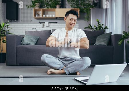 Happy Asian praktiziert Yoga zu Hause, sitzt in Lotusposition, Mann nutzt Laptop für Online-Kurse, führt einen aktiven Lebensstil Stockfoto