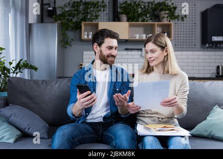 Ein Mann und eine Frau sind zu Hause glücklich, leisten Online-Zahlungen, nutzen die Telefon- und Bankanwendung, sitzen auf der Couch und halten Rechnungen Stockfoto