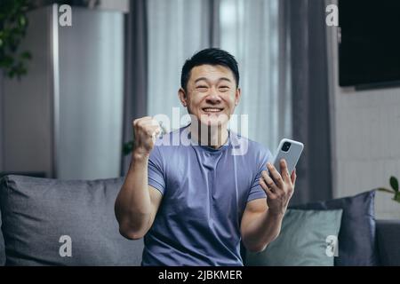 Glücklicher Mann zu Hause, erfolgreicher Sieg, online gewonnen sitzt auf der Couch, Asian schreit vor Freude und lächelt schaut auf die Kamera, hält das Telefon Stockfoto