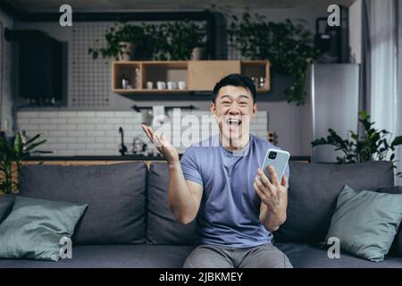 Glücklicher Mann zu Hause, erfolgreicher Sieg, online gewonnen sitzt auf der Couch, Asian schreit vor Freude und lächelt schaut auf die Kamera, hält das Telefon Stockfoto