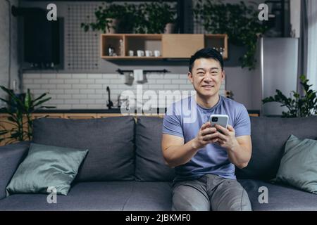 Glücklicher Mann zu Hause, erfolgreicher Sieg, online gewonnen sitzt auf der Couch, Asian schreit vor Freude und lächelt schaut auf die Kamera, hält das Telefon Stockfoto